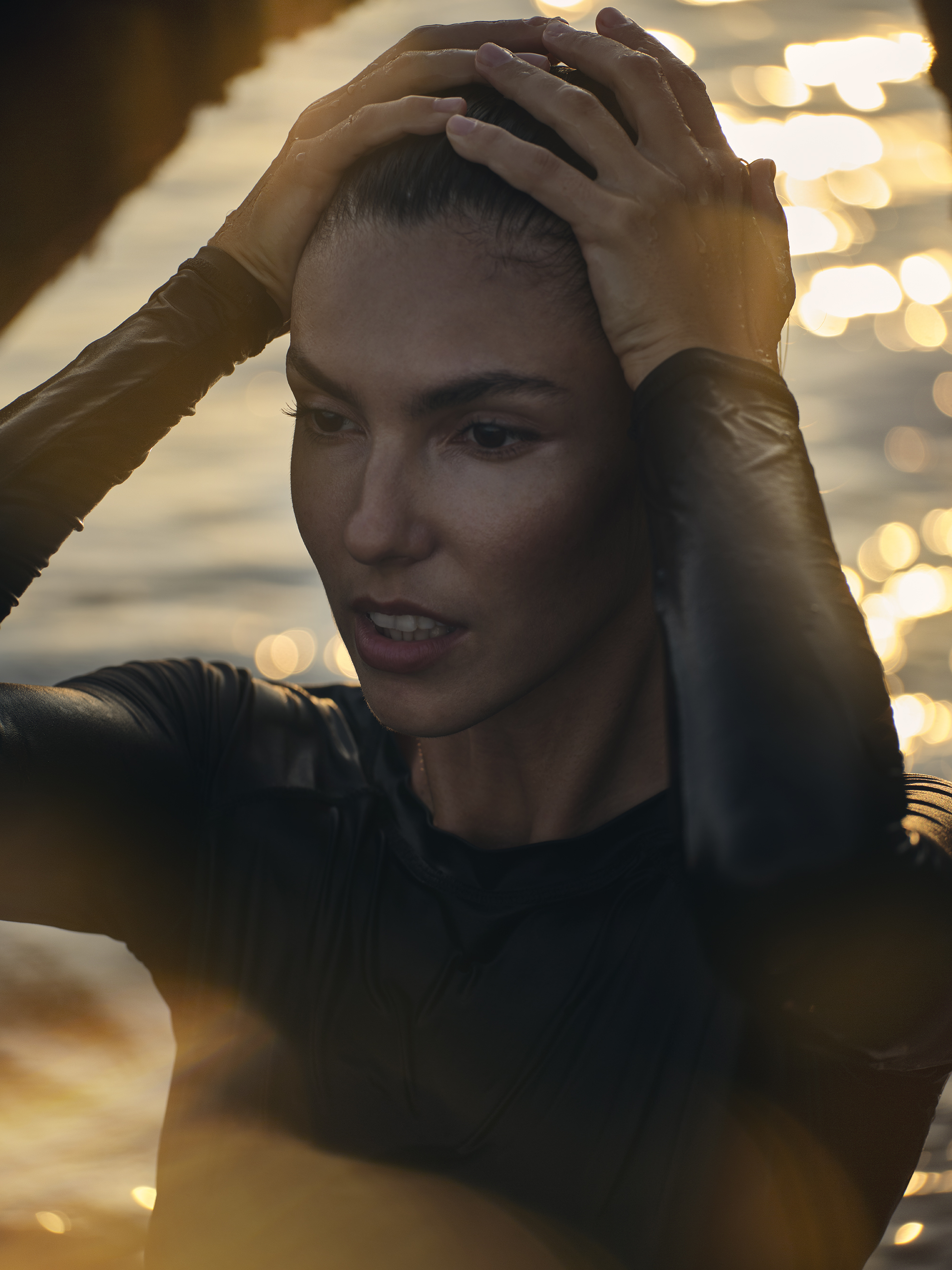 A woman in a black long-sleeved shirt stands in water, holding her head with both hands in front of a blurred golden sunset backdrop.