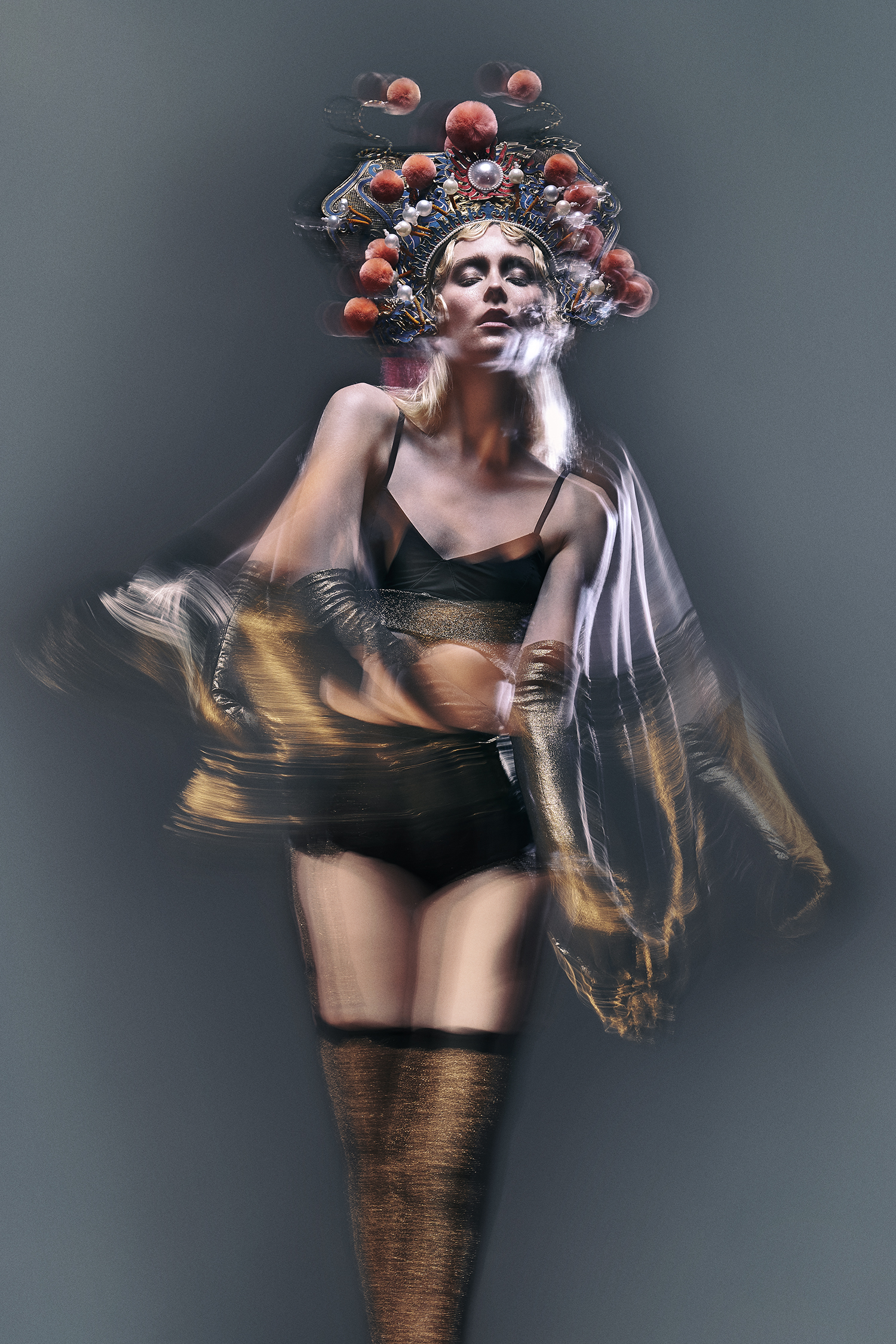 Adult female model with stage makeup and theatrical headwear poses in a studio with a blurred long-exposure effect on sheer fabric.