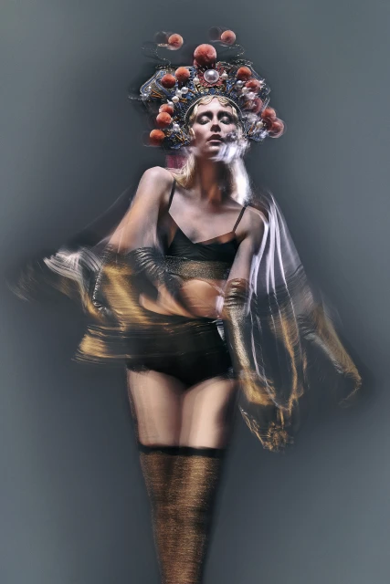 Adult female model with stage makeup and theatrical headwear poses in a studio with a blurred long-exposure effect on sheer fabric.