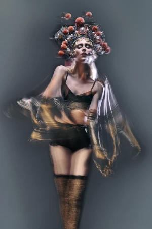 Adult female model with stage makeup and theatrical headwear poses in a studio with a blurred long-exposure effect on sheer fabric.