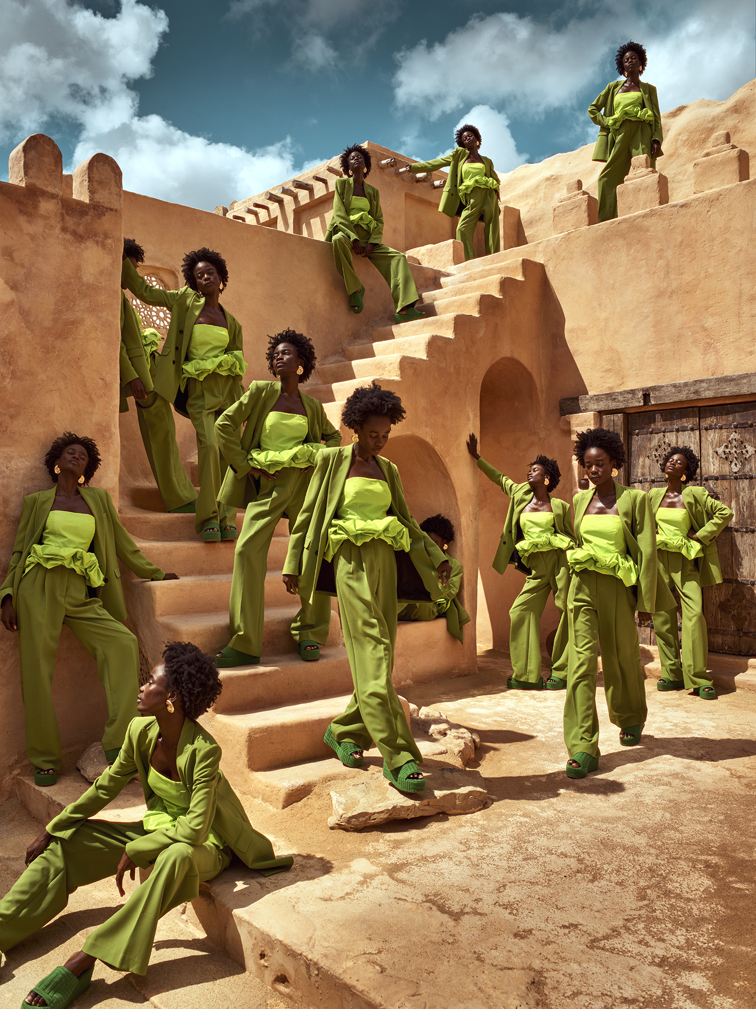 Many African American women in matching green outfits pose on tan steps under a blue sky, creating a visually striking fashion tableau.