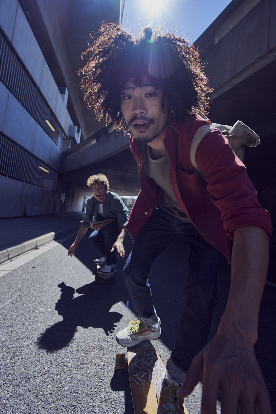 Two young men with curly hair skateboarding on asphalt road, with an urban tunnel background, bathed in sunlight. Skateboarding, asphalt, urban setting.