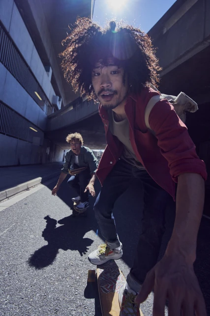 Two young men with curly hair skateboarding on asphalt road, with an urban tunnel background, bathed in sunlight. Skateboarding, asphalt, urban setting.