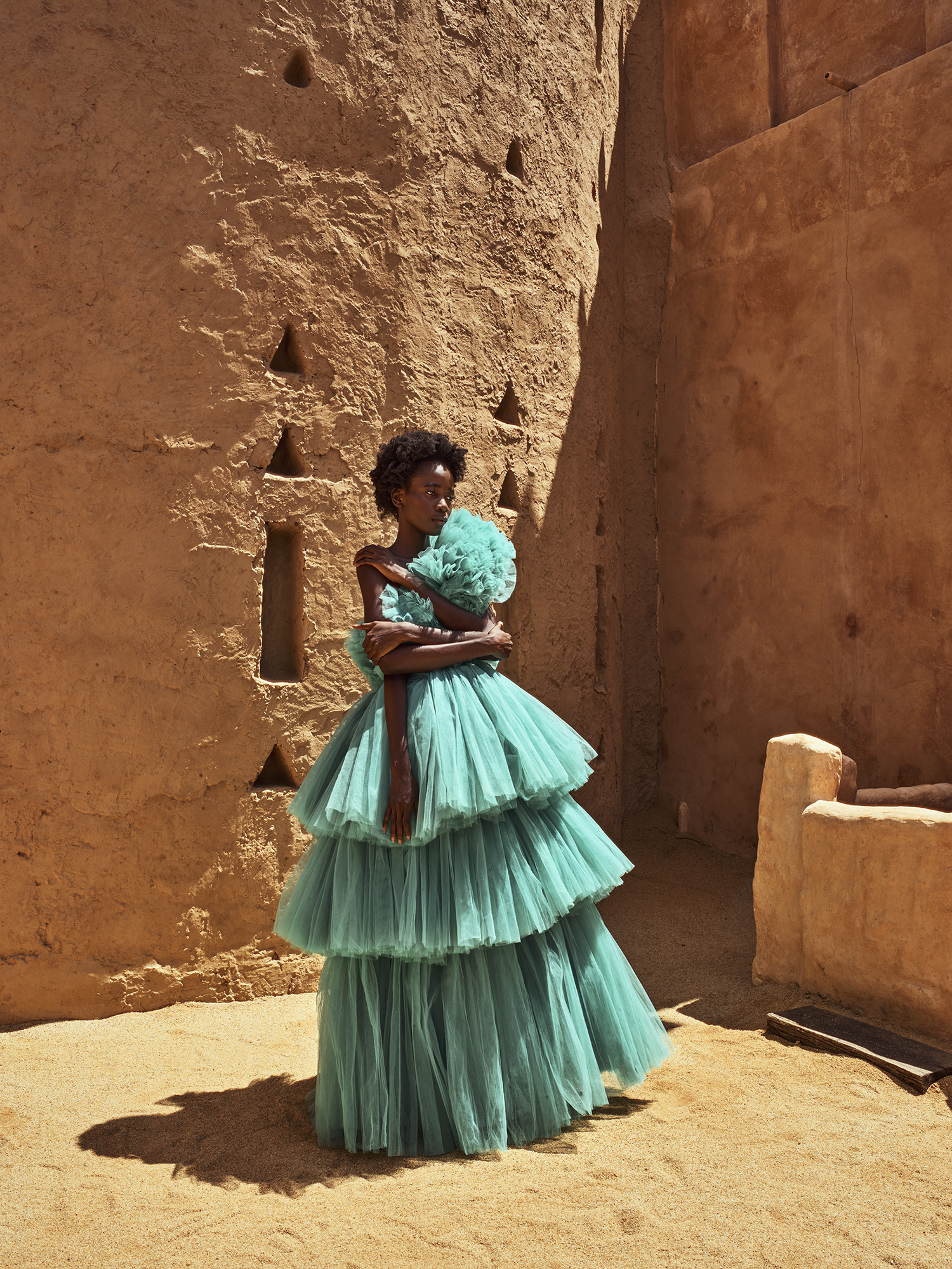 Fashion shot of a Black woman in a tiered aqua gown posing against a textured earthen wall in an outdoor setting with bright, direct sunlight.