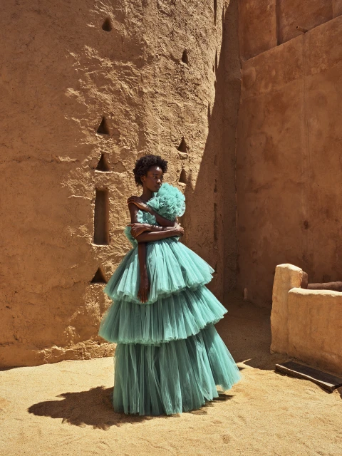 Fashion shot of a Black woman in a tiered aqua gown posing against a textured earthen wall in an outdoor setting with bright, direct sunlight.