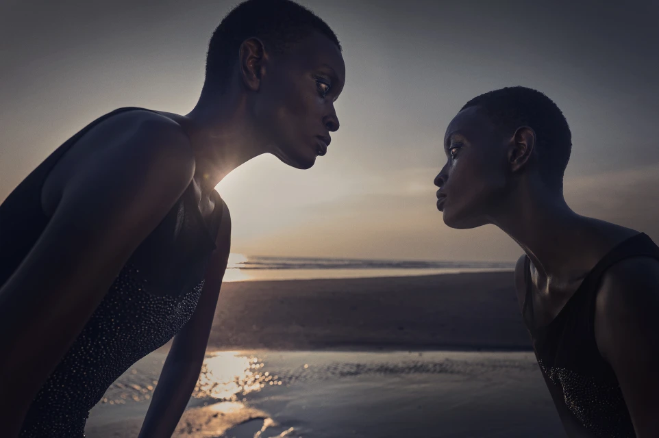 Two African female models in swimsuits face each other on a beach at sunset, creating a striking contrast against the horizon.
