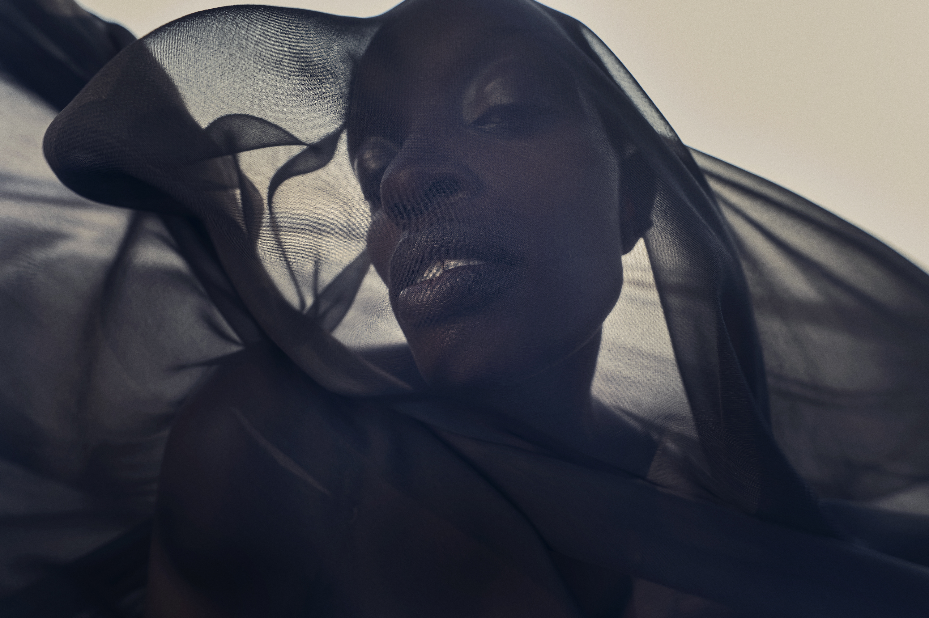 Close-up of a Black woman's face with a sheer black fabric partially covering her face and shoulders in front of a blurred background.