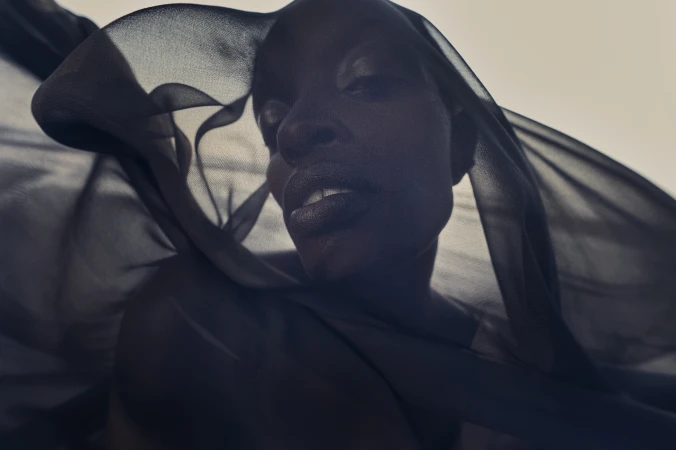 Close-up of a Black woman's face with a sheer black fabric partially covering her face and shoulders in front of a blurred background.
