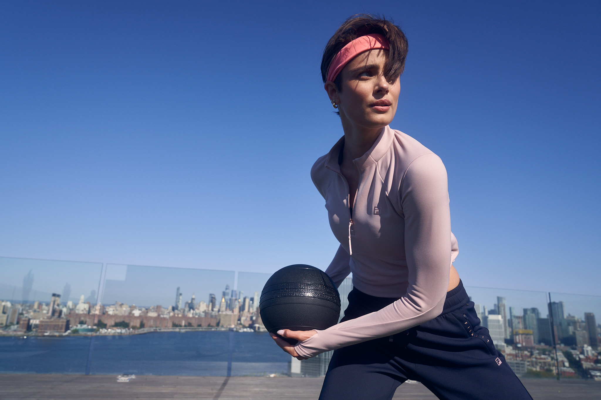 Adult female in activewear works out with a medicine ball on a rooftop with a cityscape and blue sky in the background.