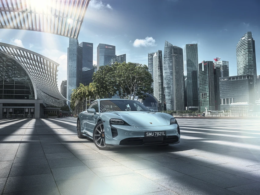 Light blue Porsche Taycan electric car parked in an urban city center with modern architecture and skyscrapers under a blue sky.