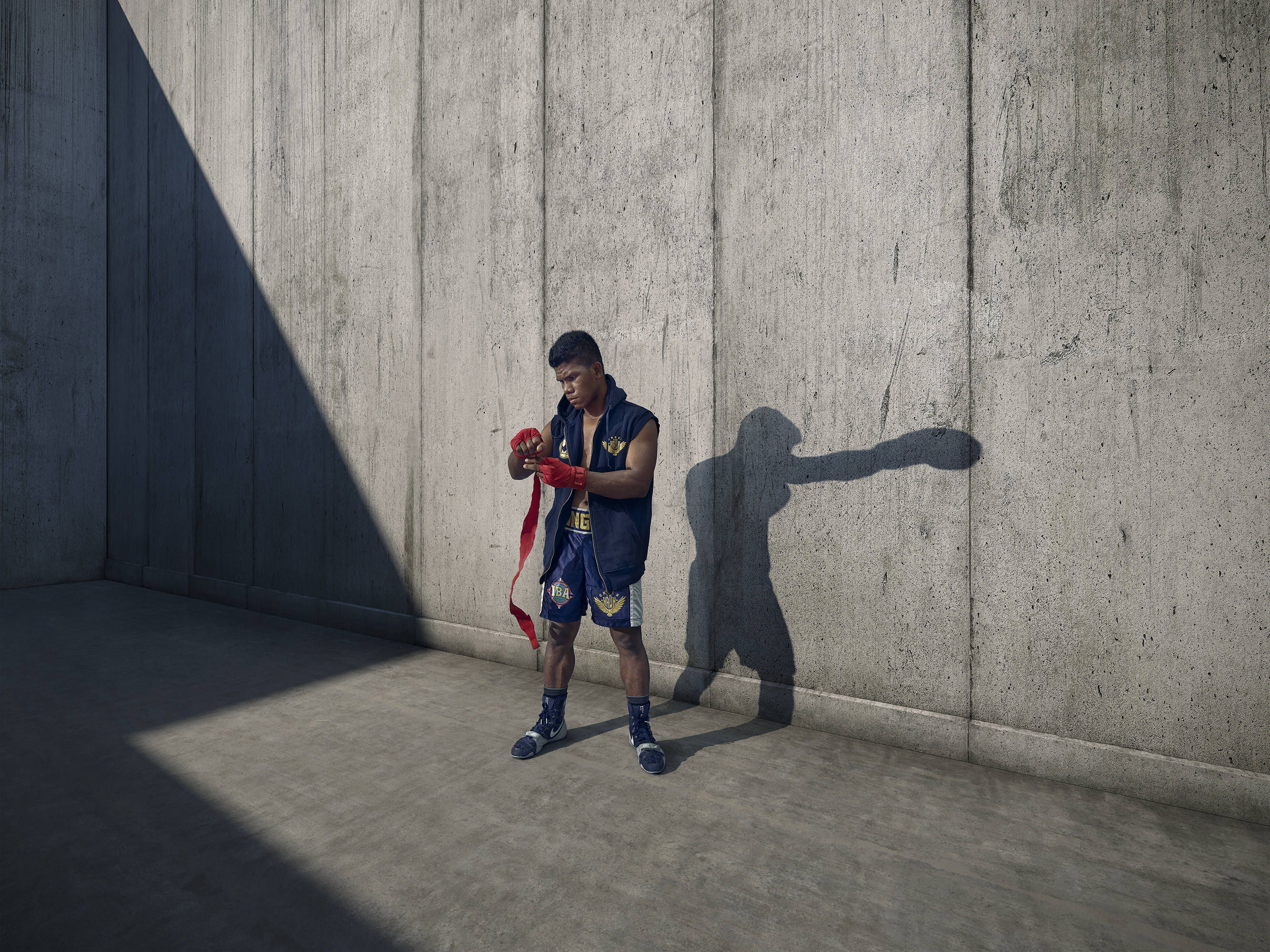 Boxer wraps hands in outdoor concrete space. Athlete prepares in blue shorts and shadow, showcasing fitness in urban setting.
