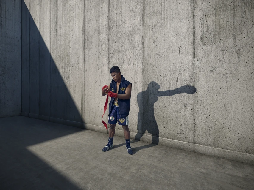 Boxer wraps hands in outdoor concrete space. Athlete prepares in blue shorts and shadow, showcasing fitness in urban setting.