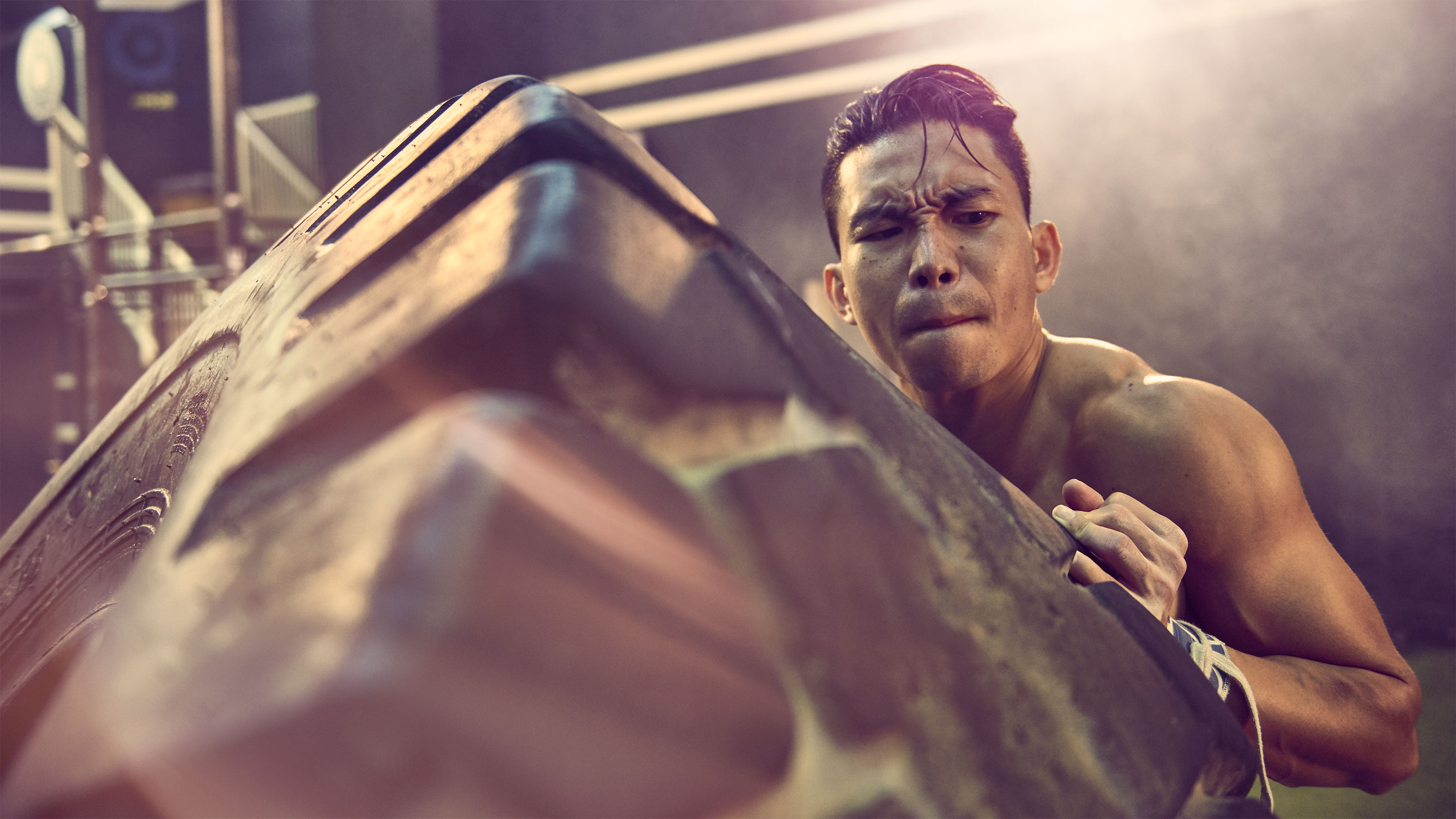 Muscular adult male lifts heavy rubber tire in an indoor gym setting, showcasing fitness and strength with dramatic lighting.