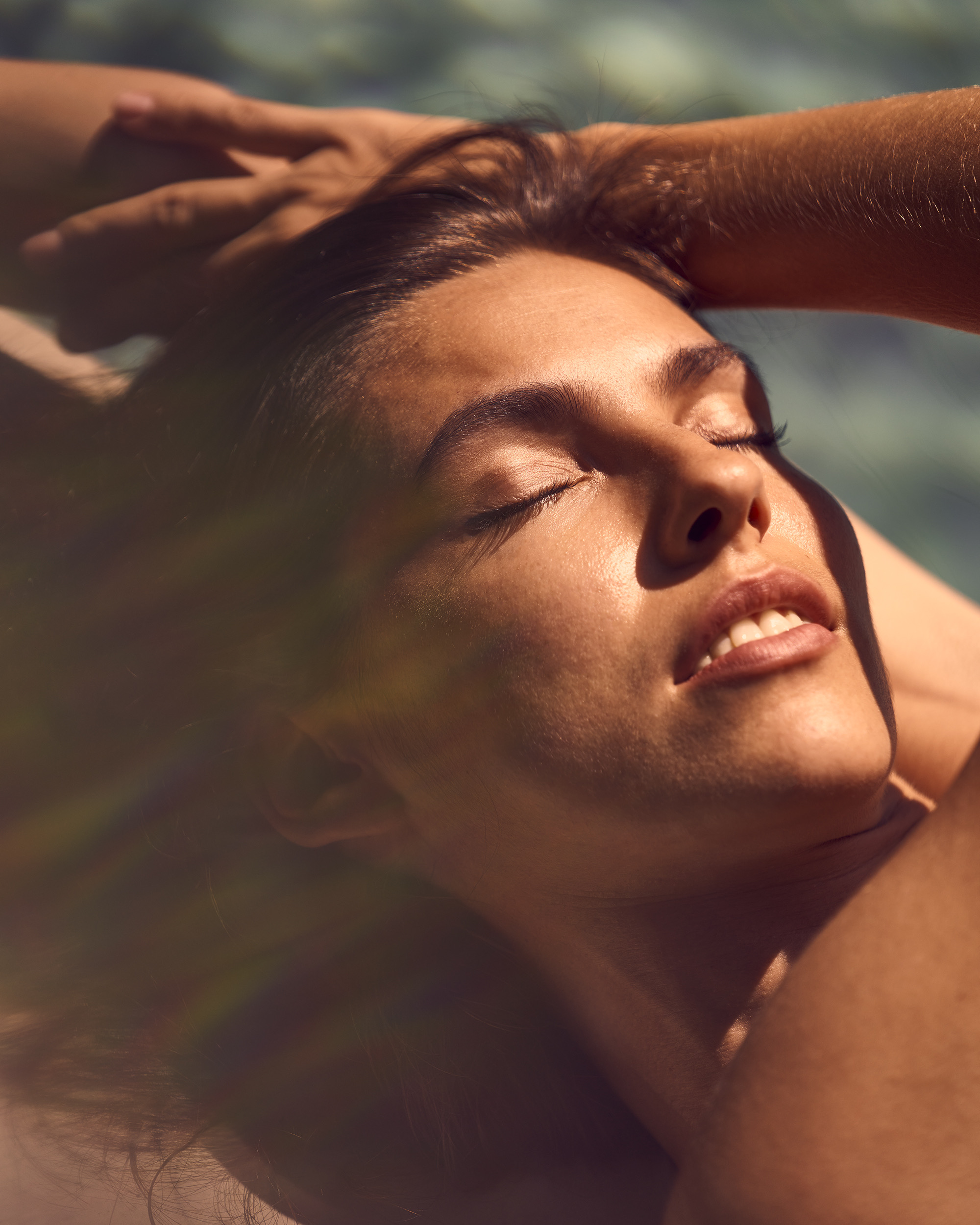 Close-up portrait of a woman resting with eyes closed and face turned toward warm direct sunlight with hands behind her head.