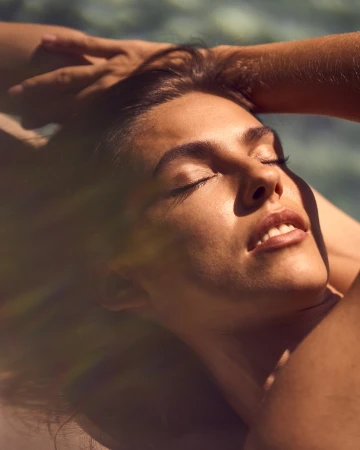 Close-up portrait of a woman resting with eyes closed and face turned toward warm direct sunlight with hands behind her head.