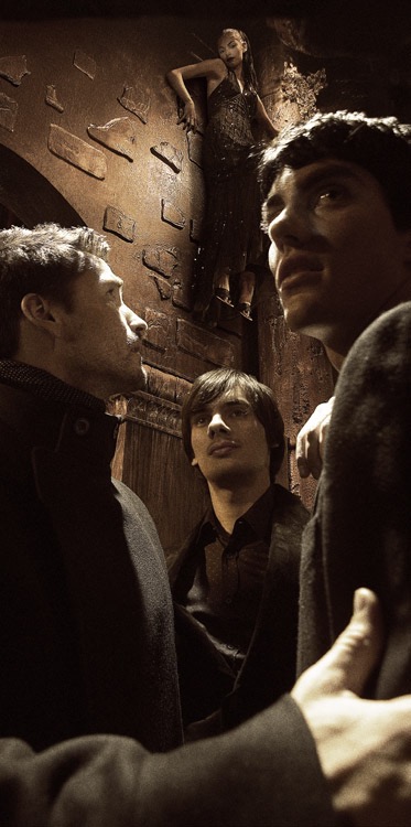 Group of four people in dim light. Three men look upwards. A woman stands above them on a brick ledge, wearing a dark, sparkly dress.