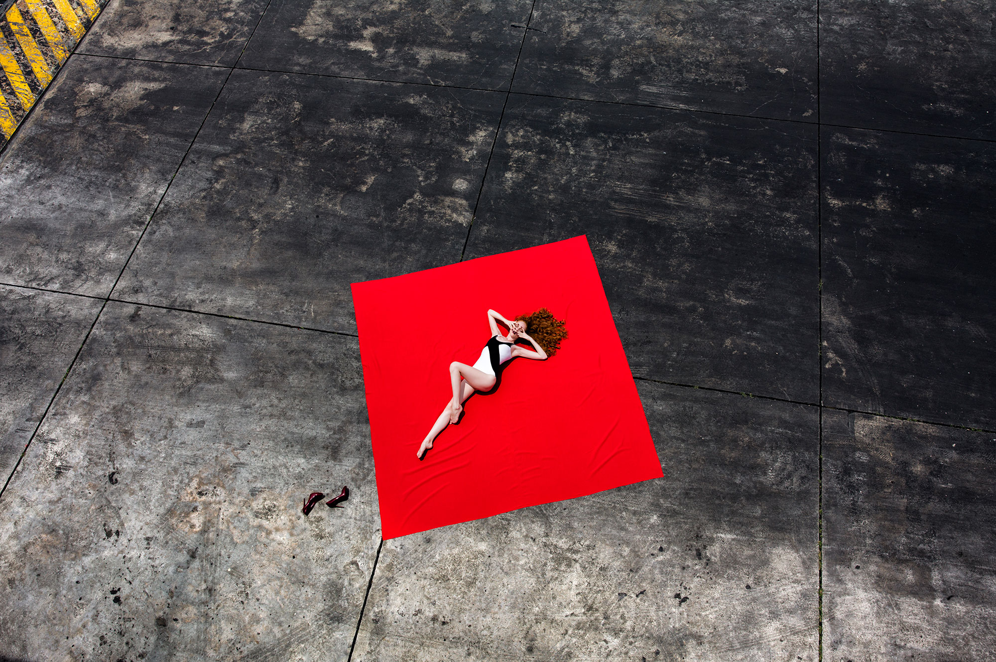 Woman in swimsuit reclining on red fabric outdoors. Aerial perspective highlights fashion, styling, and color contrast.