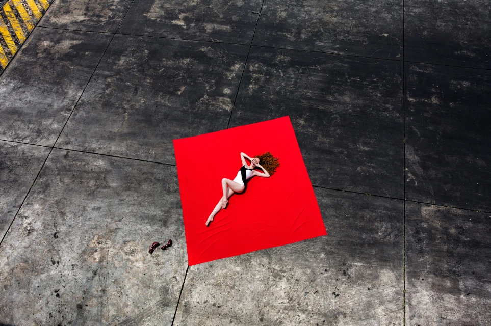 Woman in swimsuit reclining on red fabric outdoors. Aerial perspective highlights fashion, styling, and color contrast.