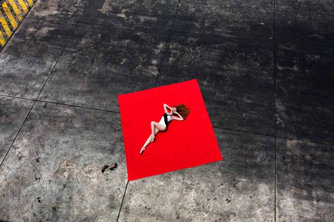 Woman in swimsuit reclining on red fabric outdoors. Aerial perspective highlights fashion, styling, and color contrast.