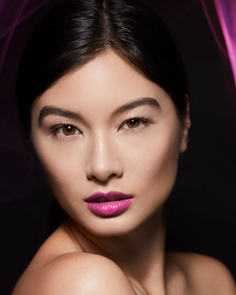 Close-up portrait of a young Asian woman with dark hair and pink lipstick against a dark background with pink lighting accents.
