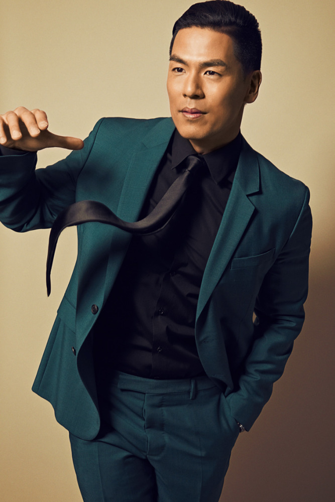 Portrait of an Asian man in a green suit and black shirt, looking to the side while holding his tie, against a neutral background.