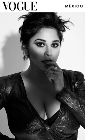 Black and white portrait of a woman with dark hair wearing a shimmery dress, posing in front of a white background with Vogue logo.