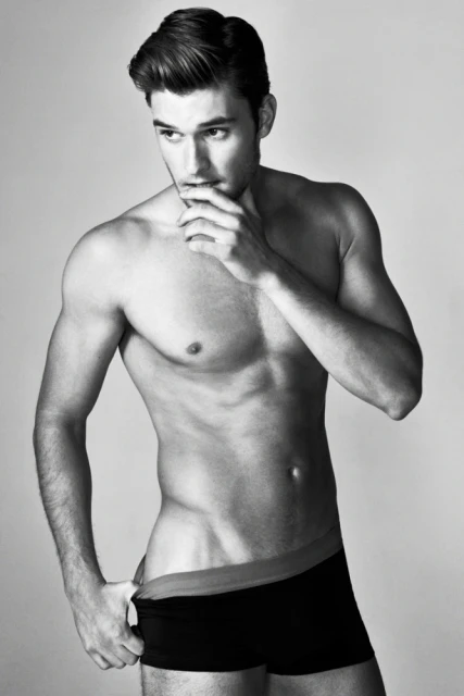 Black-and-white studio portrait of a muscular adult male in underwear, hand in waistband, looking off-camera with thoughtful expression.