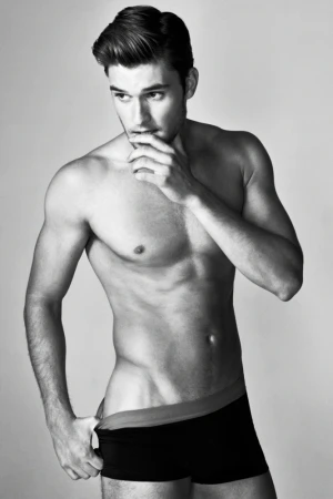 Black-and-white studio portrait of a muscular adult male in underwear, hand in waistband, looking off-camera with thoughtful expression.