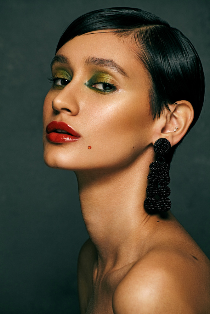 Close-up portrait of a woman with short black hair, red lipstick, and green eyeshadow against a dark grey background in a beauty shot.