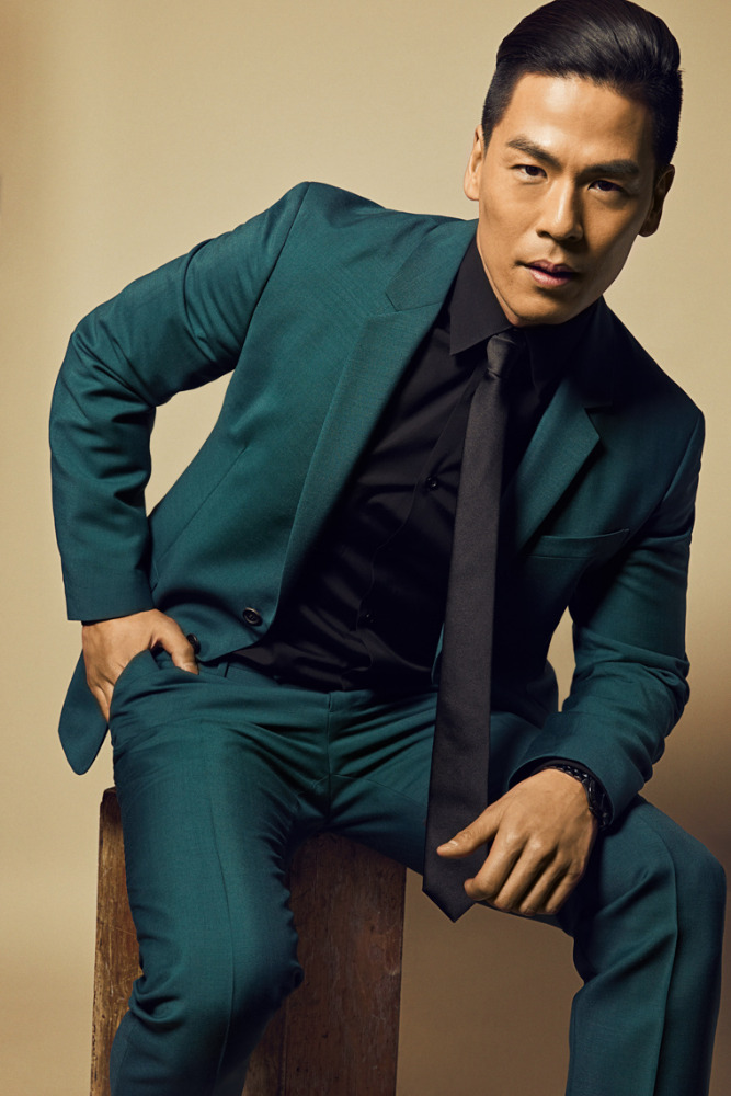 Portrait of a handsome adult male in a teal suit sitting on a wooden block in front of a tan backdrop, looking directly at the camera.