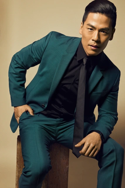 Portrait of a handsome adult male in a teal suit sitting on a wooden block in front of a tan backdrop, looking directly at the camera.