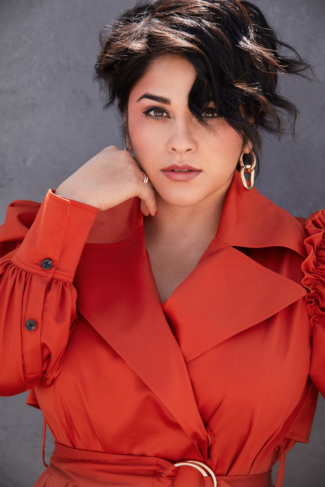 Close-up portrait of a woman with short dark hair in a red trench coat, posing against a grey backdrop with gold earrings and belt.