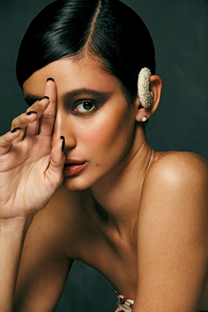 Close-up portrait of a woman with dark hair and smokey eye makeup, wearing pearl ear jewelry and covering half of her face with her hand.