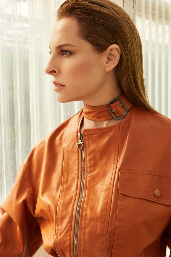 A woman poses indoors wearing a leather jacket with a choker. Her hair is slicked back, and she looks off to the side near a window.