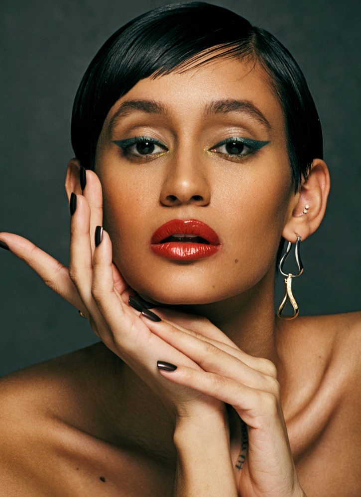 Close-up portrait of a striking brunette adult female with bold makeup, geometric earrings, and polished black nails against a gray backdrop.