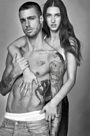 Black and white studio portrait of a man and a woman posing together. The man is shirtless and tattooed, with the woman behind him.