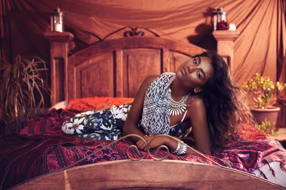 Attractive woman with brown skin poses on a bed with patterned blankets, adorned with jewelry and a white patterned shawl in a warm-toned interior.