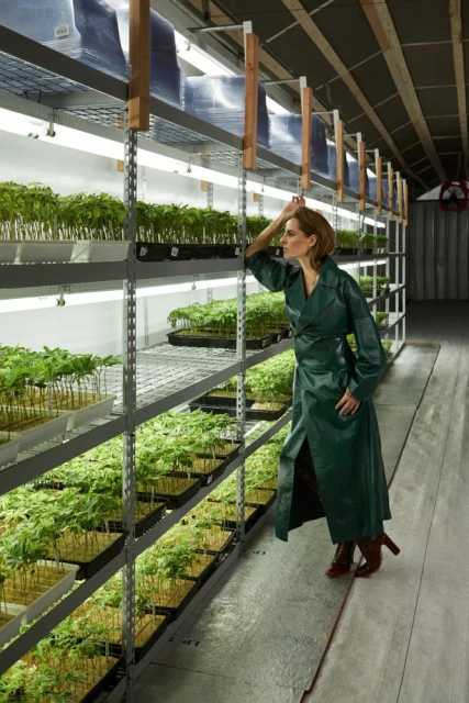 Woman in green coat in a greenhouse filled with plants on shelves, looking at greenery with neutral expression in indoor setting.