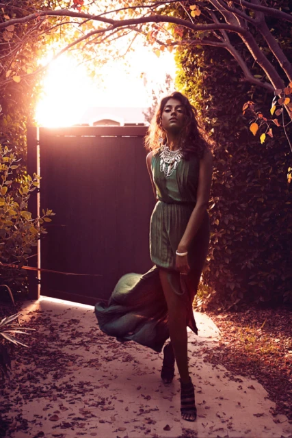 Woman in green dress walking through sunlit garden gate. She wears statement necklace and sandals. Lush foliage frames the scene.