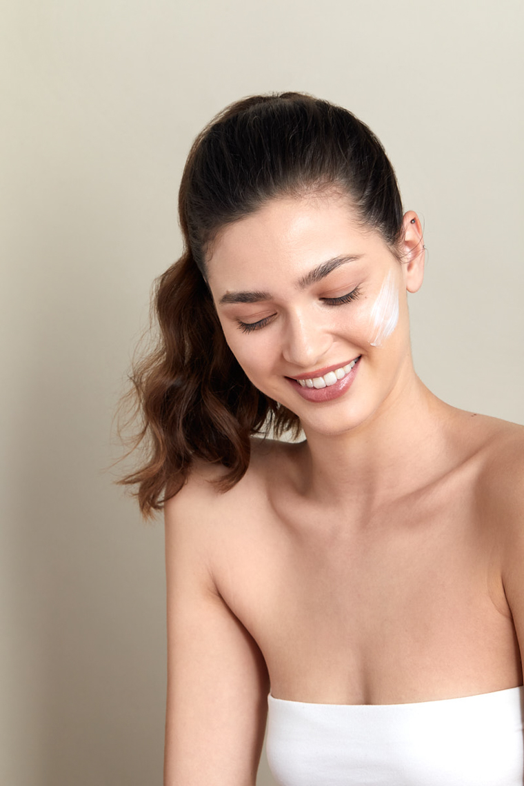 Smiling woman with clear skin and cream on her face. Beauty shot against a neutral backdrop, showcasing skincare and feminine beauty.