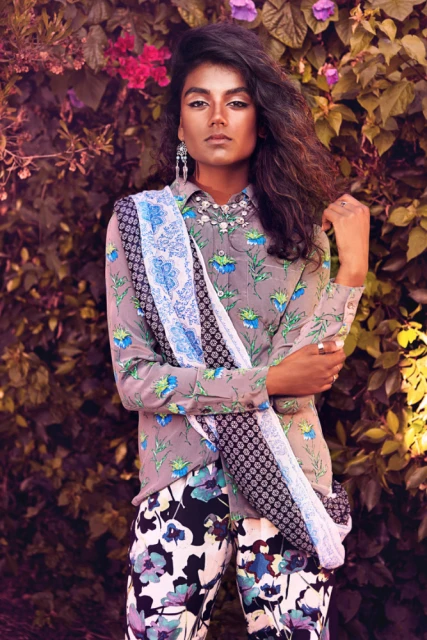 Fashion portrait of a woman in floral print top and pants, standing in front of flowering shrubs, looking at the camera with neutral expression.