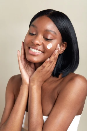 Close-up portrait of a smiling Black woman with a dab of white cream on her cheek, holding her face with both hands
