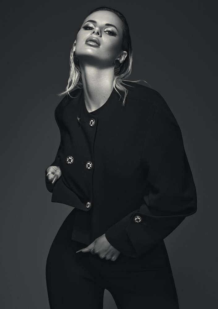 Monochromatic portrait of an adult female posing in a black jacket with silver buttons and matching pants against a grey backdrop.