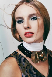 Close-up portrait of a woman with red hair, bold graphic white eyeliner, and a large black beaded necklace with a gold pendant.