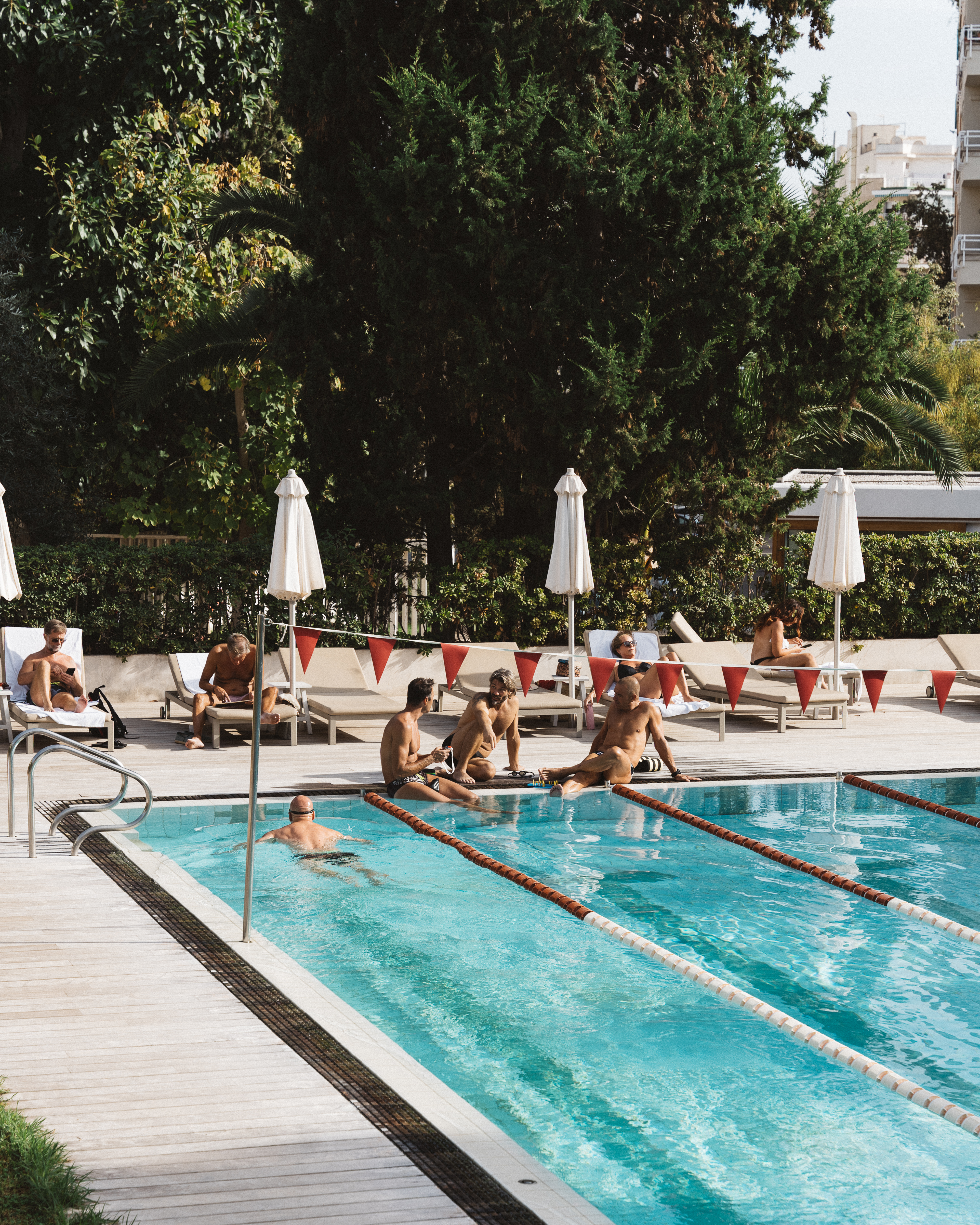 Outdoor swimming pool scene featuring a group of men sunbathing and relaxing on lounge chairs under white umbrellas, surrounded by green trees.
