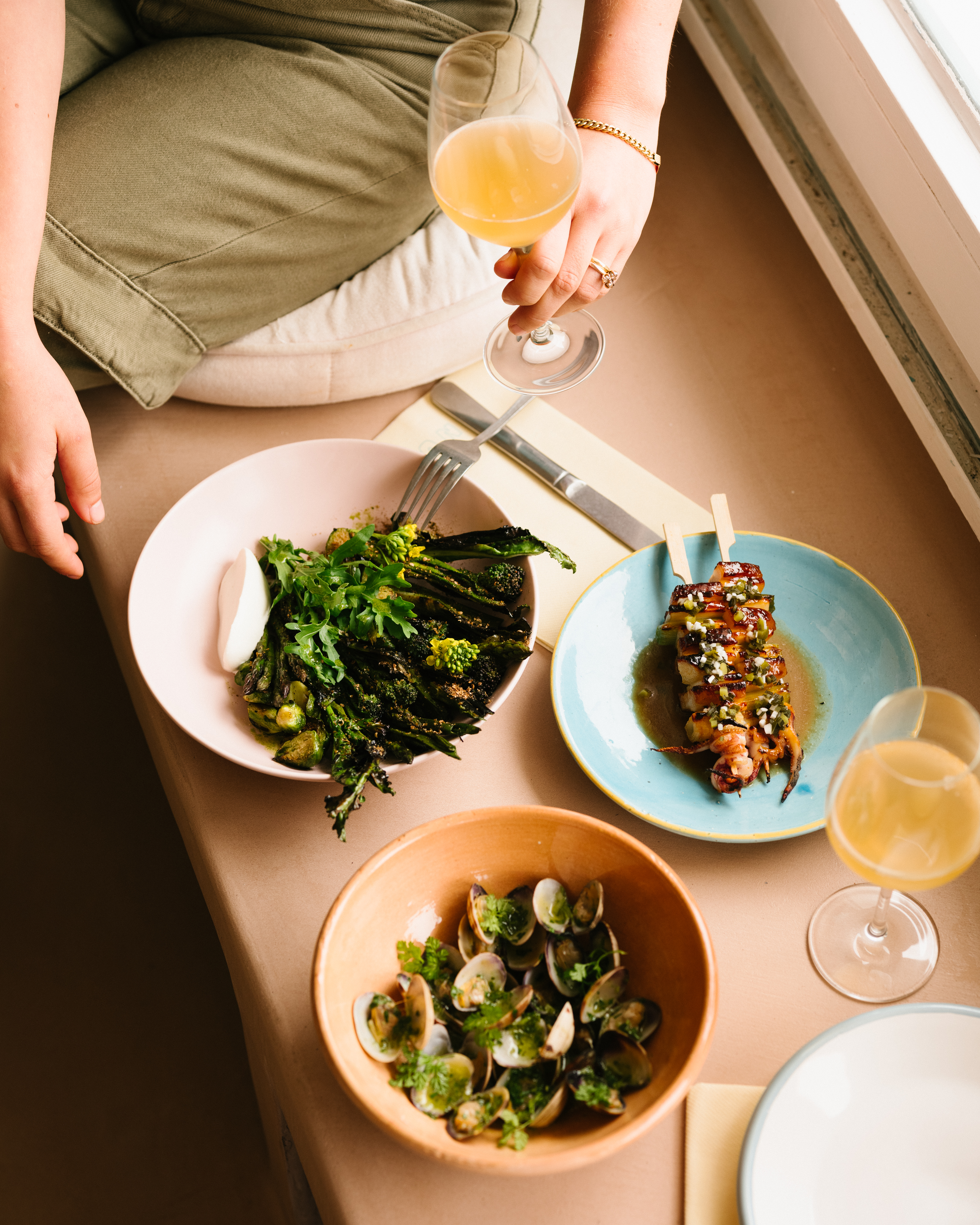 Overhead shot of a person holding a wine glass while sitting near a table with plates of food, including clams and vegetables.