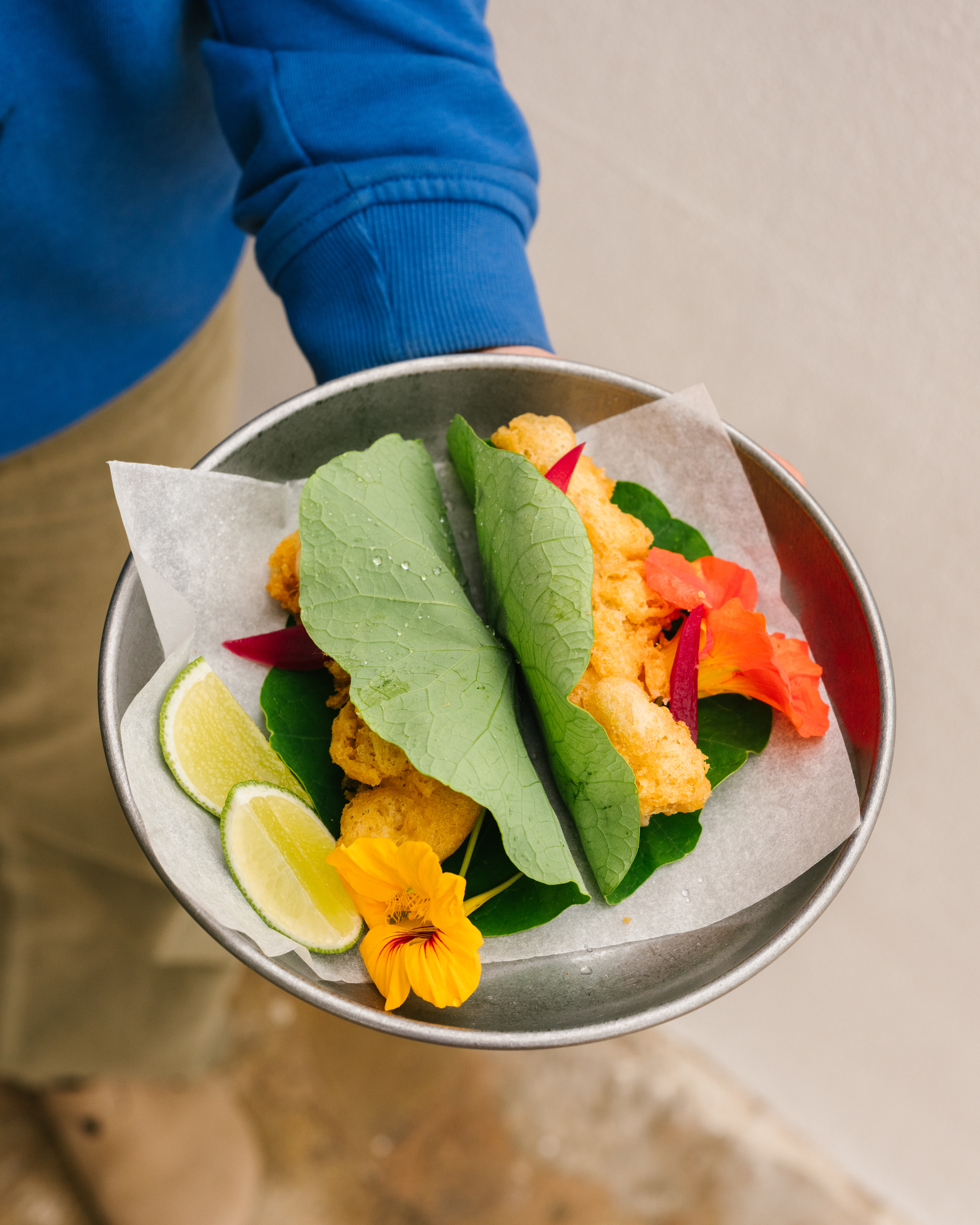 Person holding a metal plate with fried food, green leaves, lime slices, and edible orange flowers, on a neutral background.