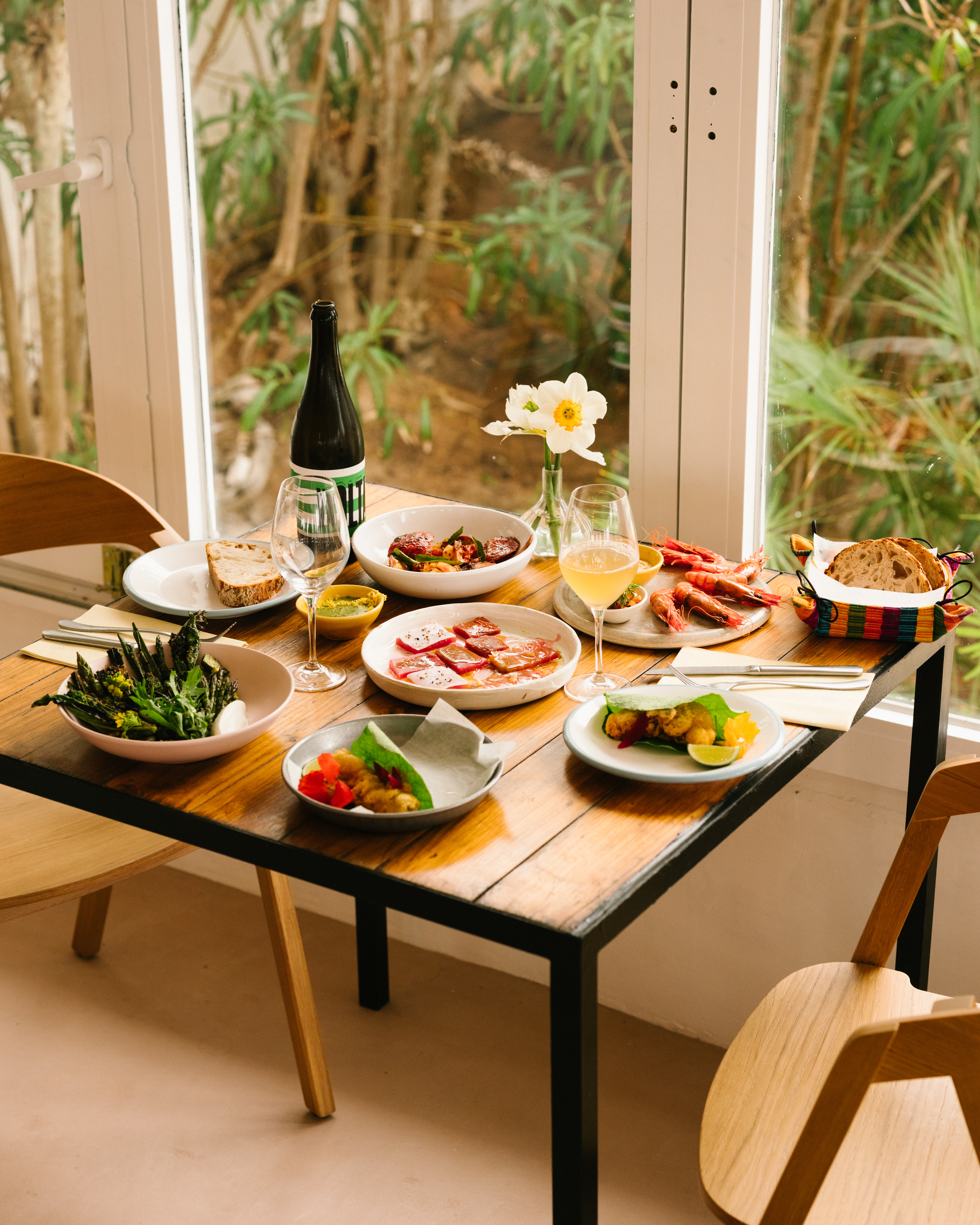 Overhead shot of a wood table laden with plates of varied food, wine, and flowers, showcasing a dining setup next to a window.