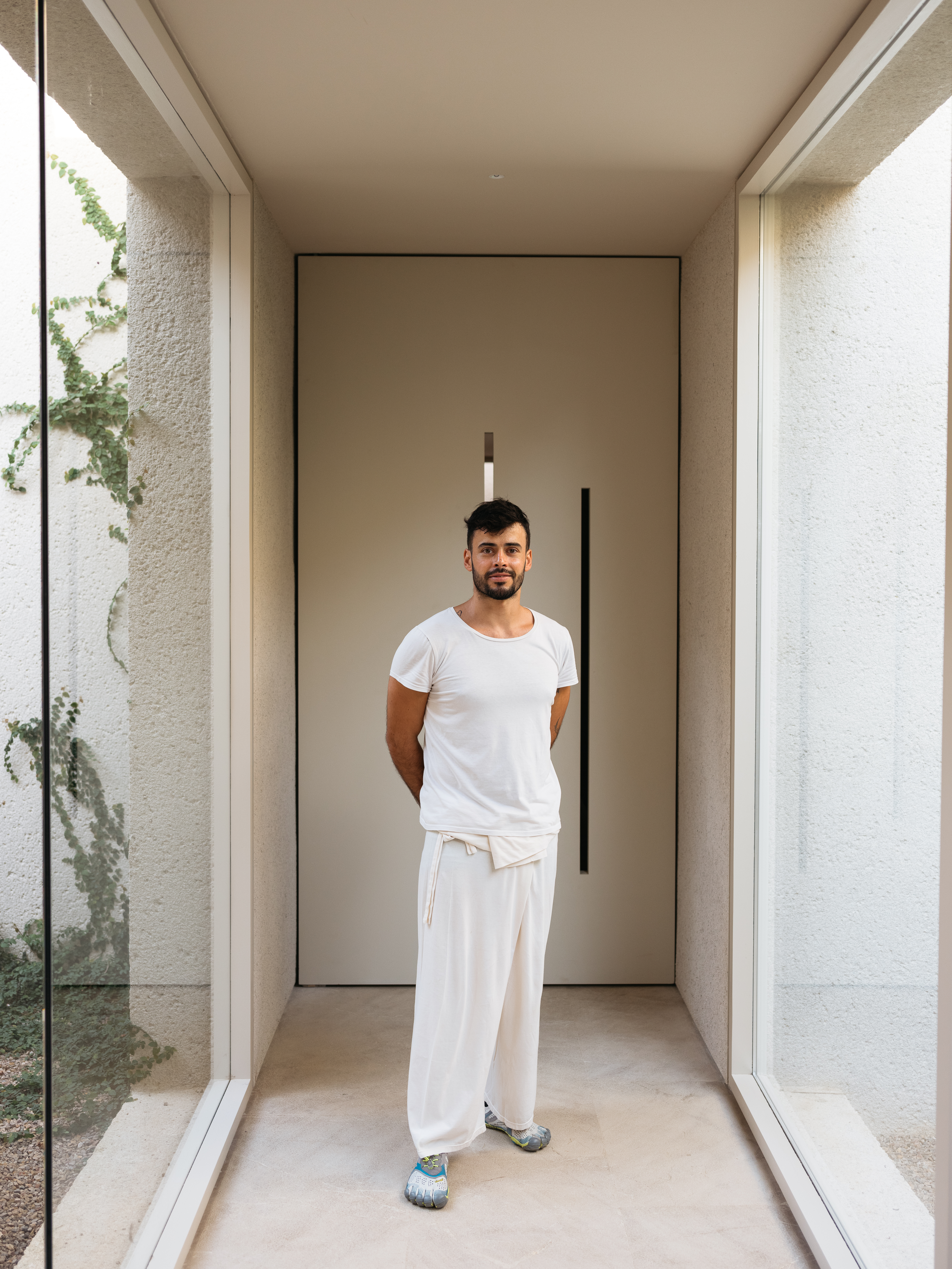 Full shot of a man in a white shirt and pants standing inside a brightly lit entryway of a modern house. Neutral color palette.