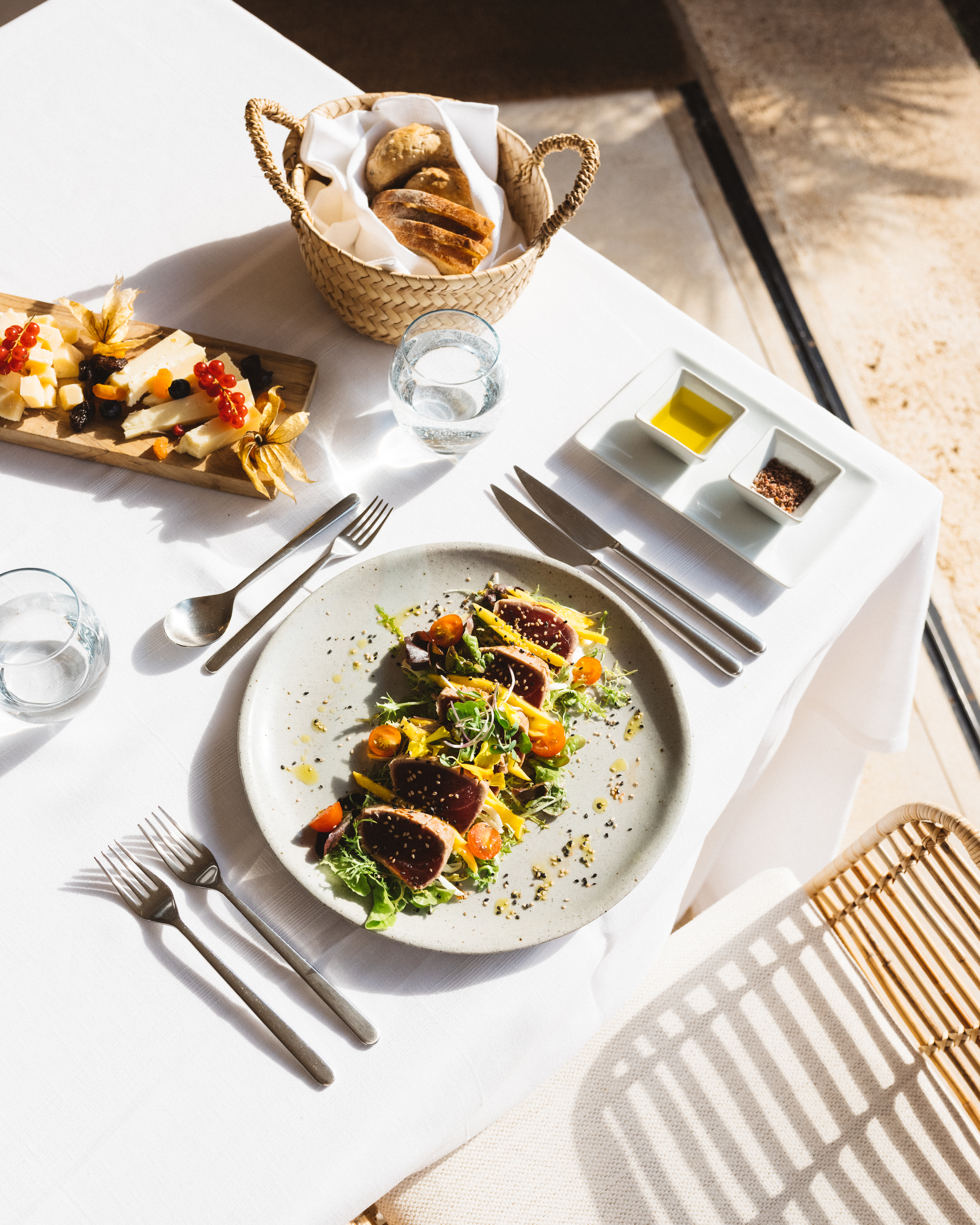 Overhead shot of tuna salad on a plate, with cheese, bread, water, and silverware, arranged on a white table with wicker chair.